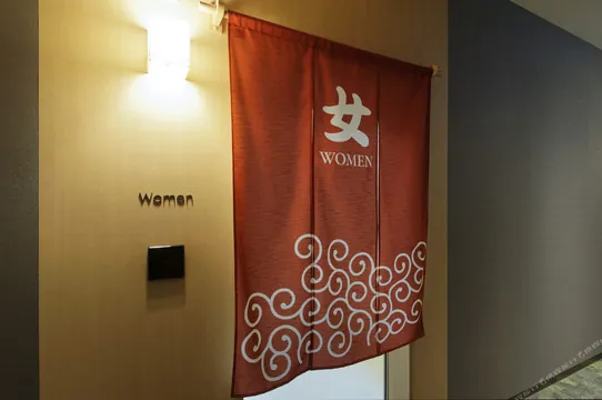 여탕 입구 노렌 (일본 전통 천 가림막). '女 WOMEN' 사인 + 아이누 문양 + 우드톤이 진짜 일본 분위기 살려줘요.